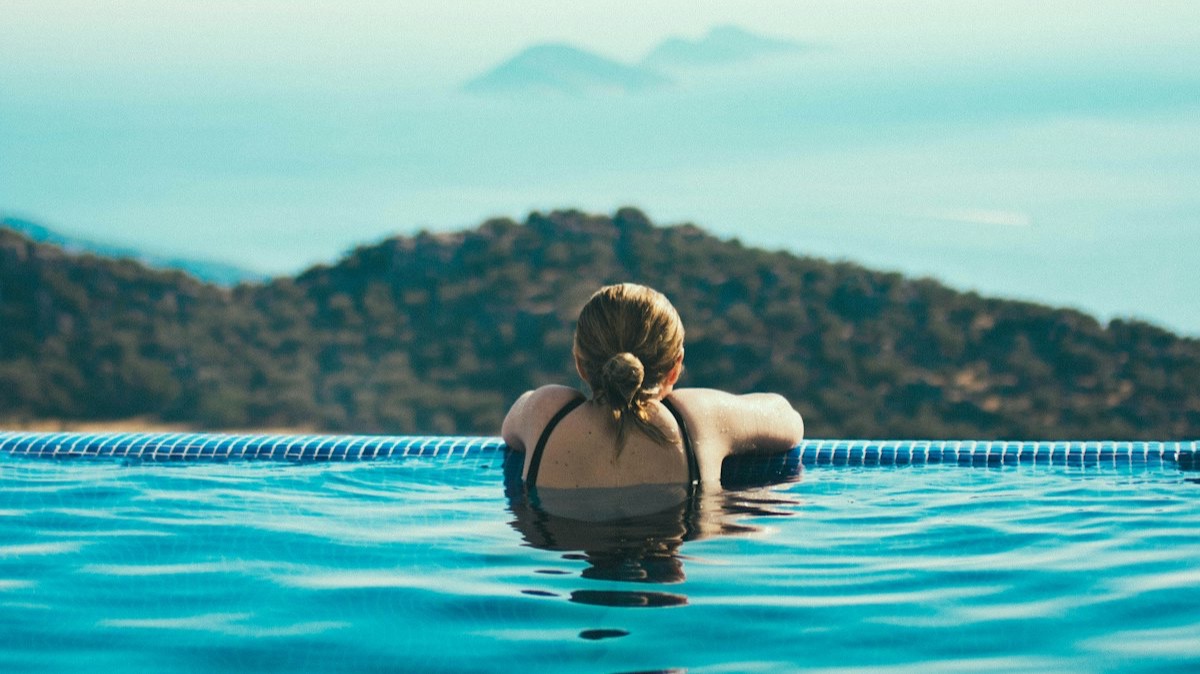 Une femme dans une piscine à débordement, face à la mer et à une côte montagneuse