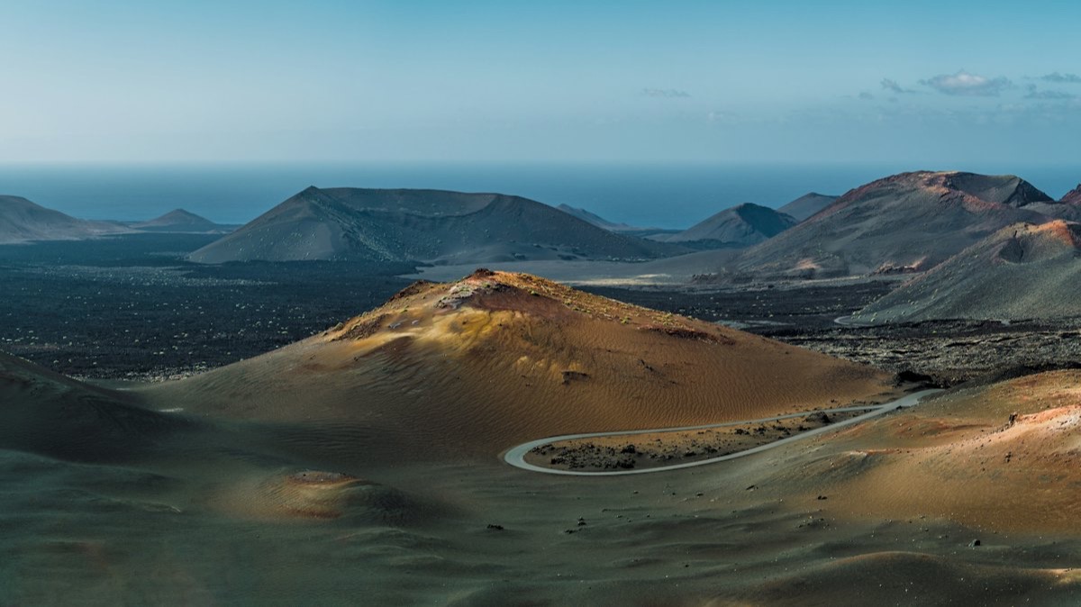 Des cônes volcaniques ocre et noirs à Lanzarote, traversés par une route sinueuse, avec l'océan à l'horizon