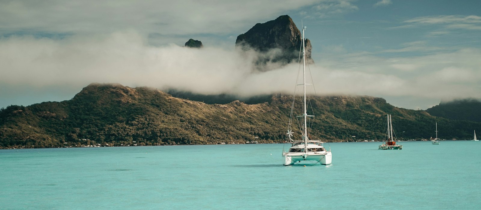 Des catamarans au mouillage dans le lagon turquoise de Bora Bora, au pied du mont Otemanu pris dans les nuages