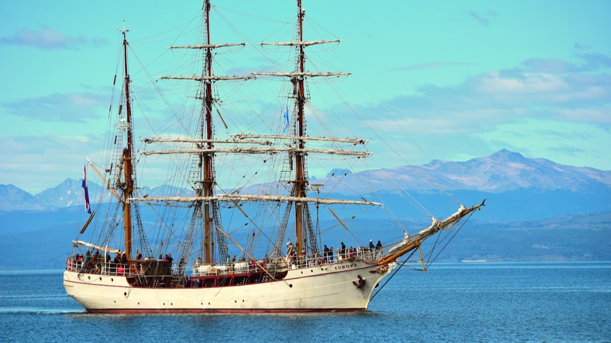 Un grand voilier à trois mâts, toutes voiles carguées, sur une mer bleue devant des montagnes