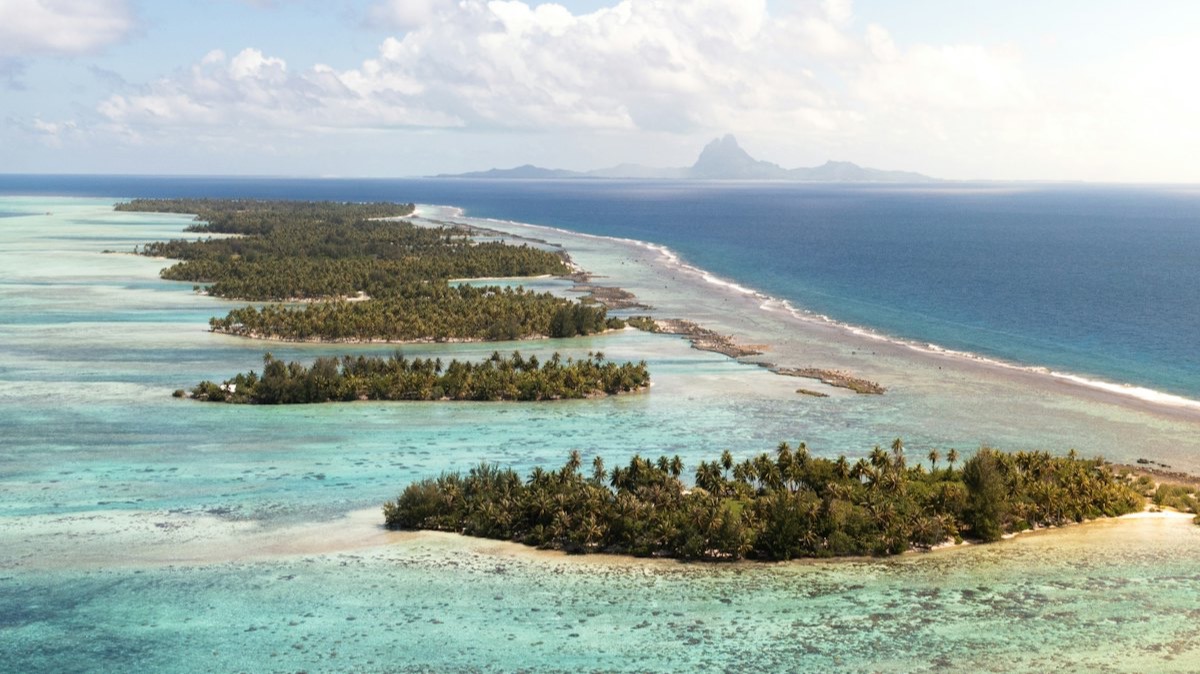Des motus couverts de cocotiers sur le récif de Taha'a, avec la silhouette de Bora Bora à l'horizon