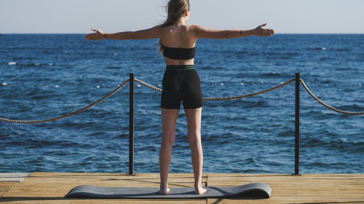 Une femme, bras tendus, sur un tapis de yoga posé sur un ponton en bois face à la mer