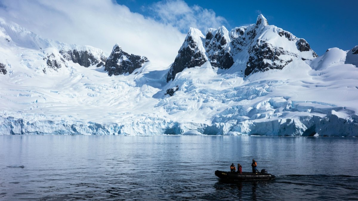 Un zodiac navigue entre des glaciers et des montagnes enneigées, sous un ciel bleu