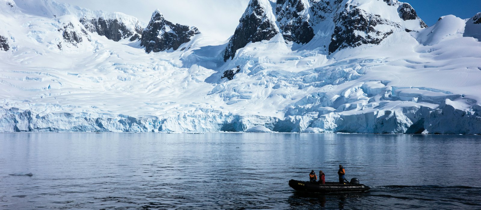 Un zodiac et ses passagers devant un glacier et des sommets enneigés, en Antarctique