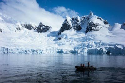 Un zodiac et ses passagers devant un glacier et des sommets enneigés, en Antarctique
