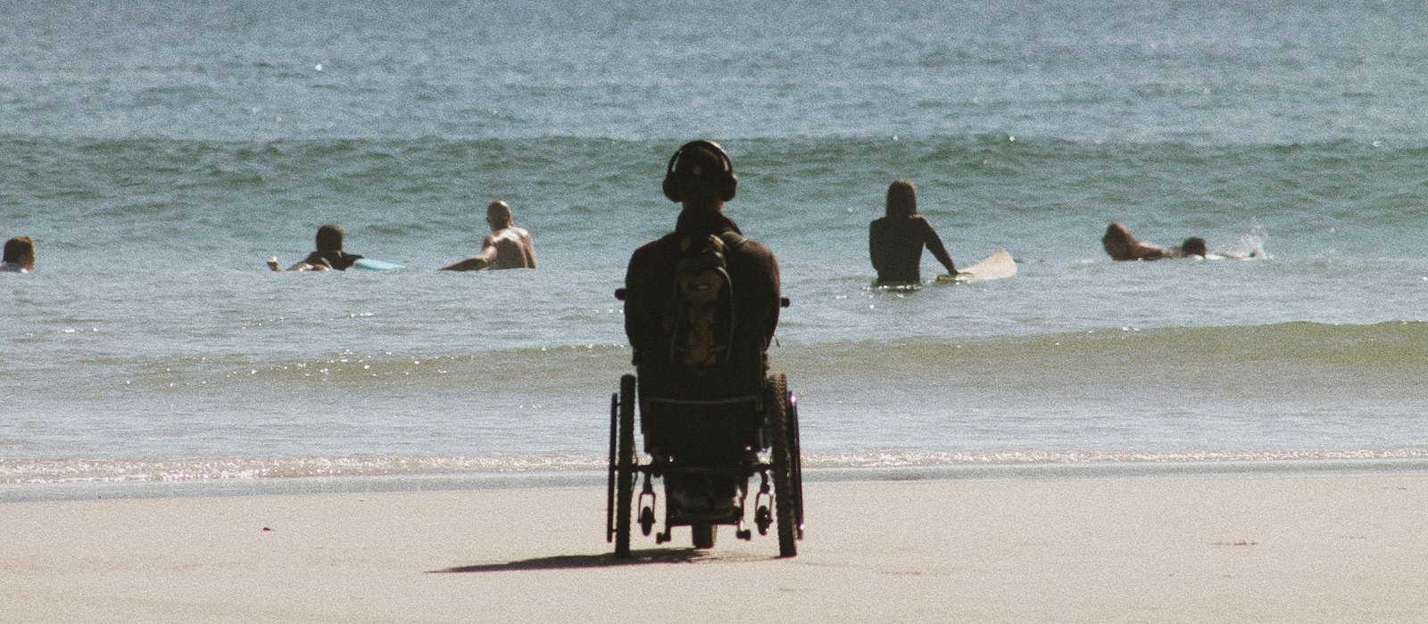 Une personne en fauteuil roulant, vue de dos, face à la mer sur une plage où nagent des surfeurs