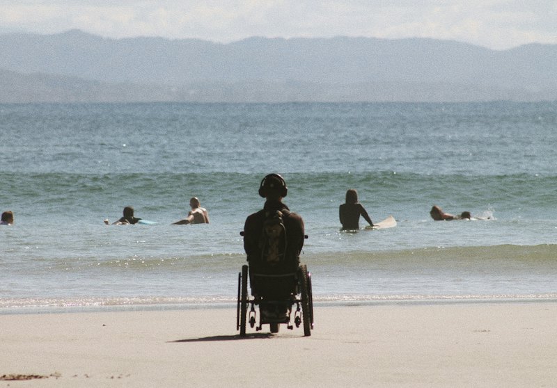 Une personne en fauteuil roulant, vue de dos, face à la mer sur une plage où nagent des surfeurs