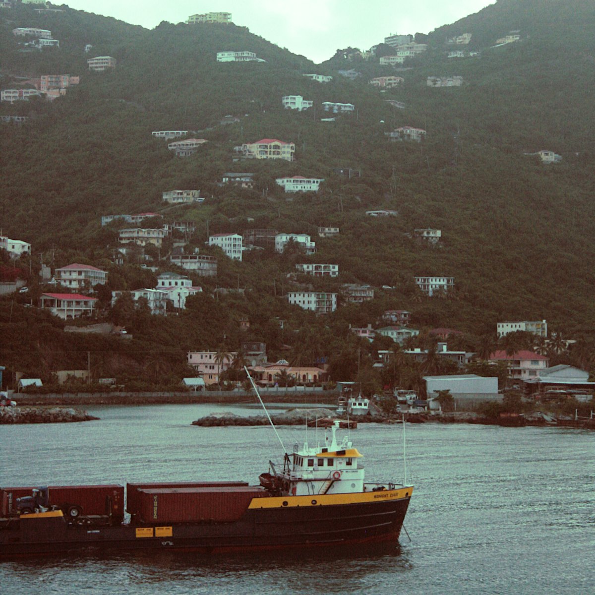 Petit cargo à conteneurs mouillant devant un village accroché au flanc d'une île