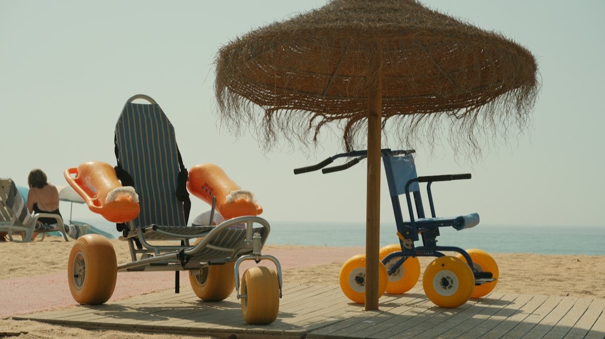 Deux fauteuils de plage à gros pneus jaunes sous un parasol de paille, sur un ponton en bois posé sur le sable